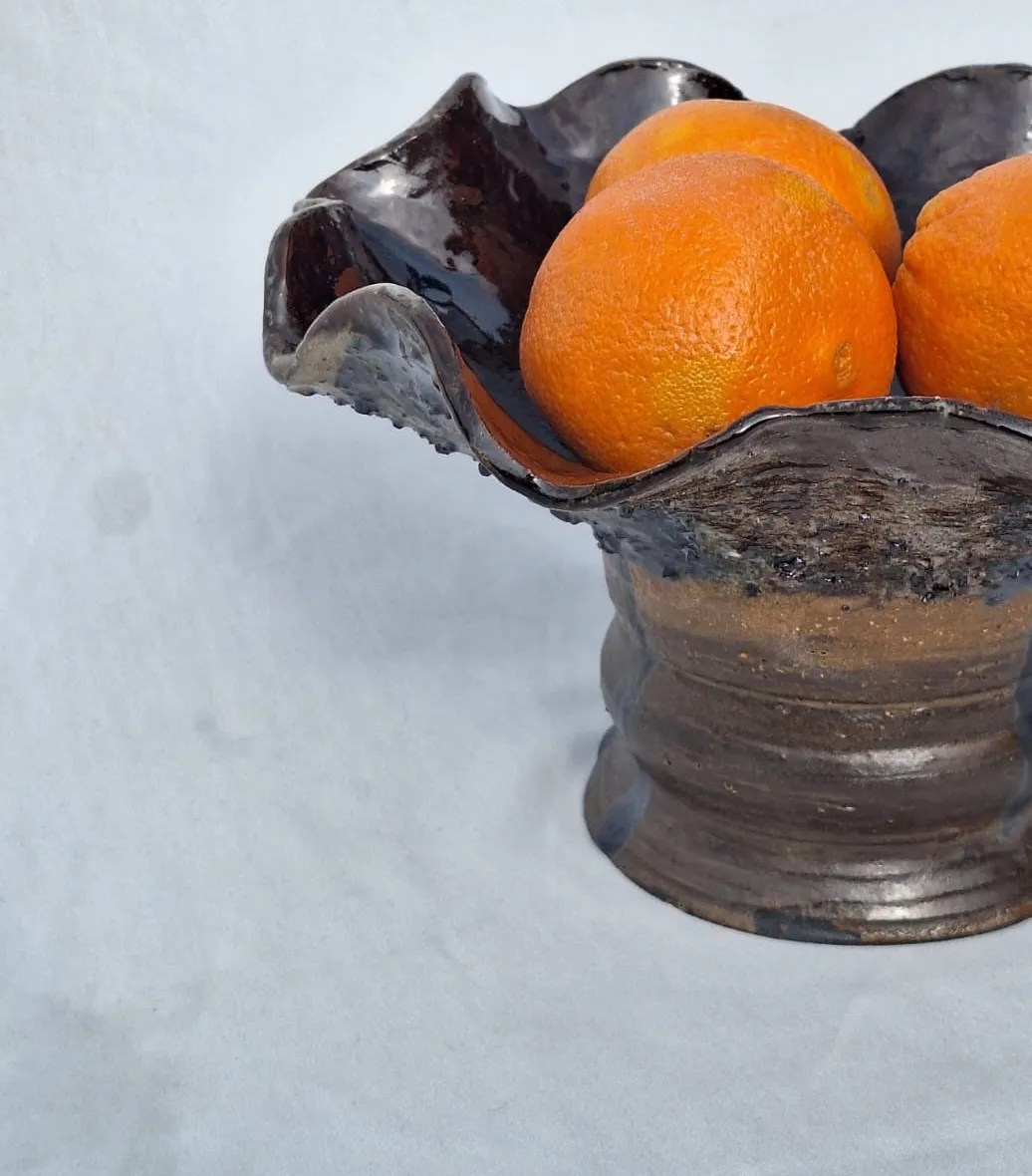Hand-built pedestal bowl with a ruffled dark glaze, holding oranges